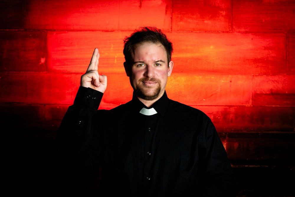 Man in black clergy shirt pointing finger up against red wall background.