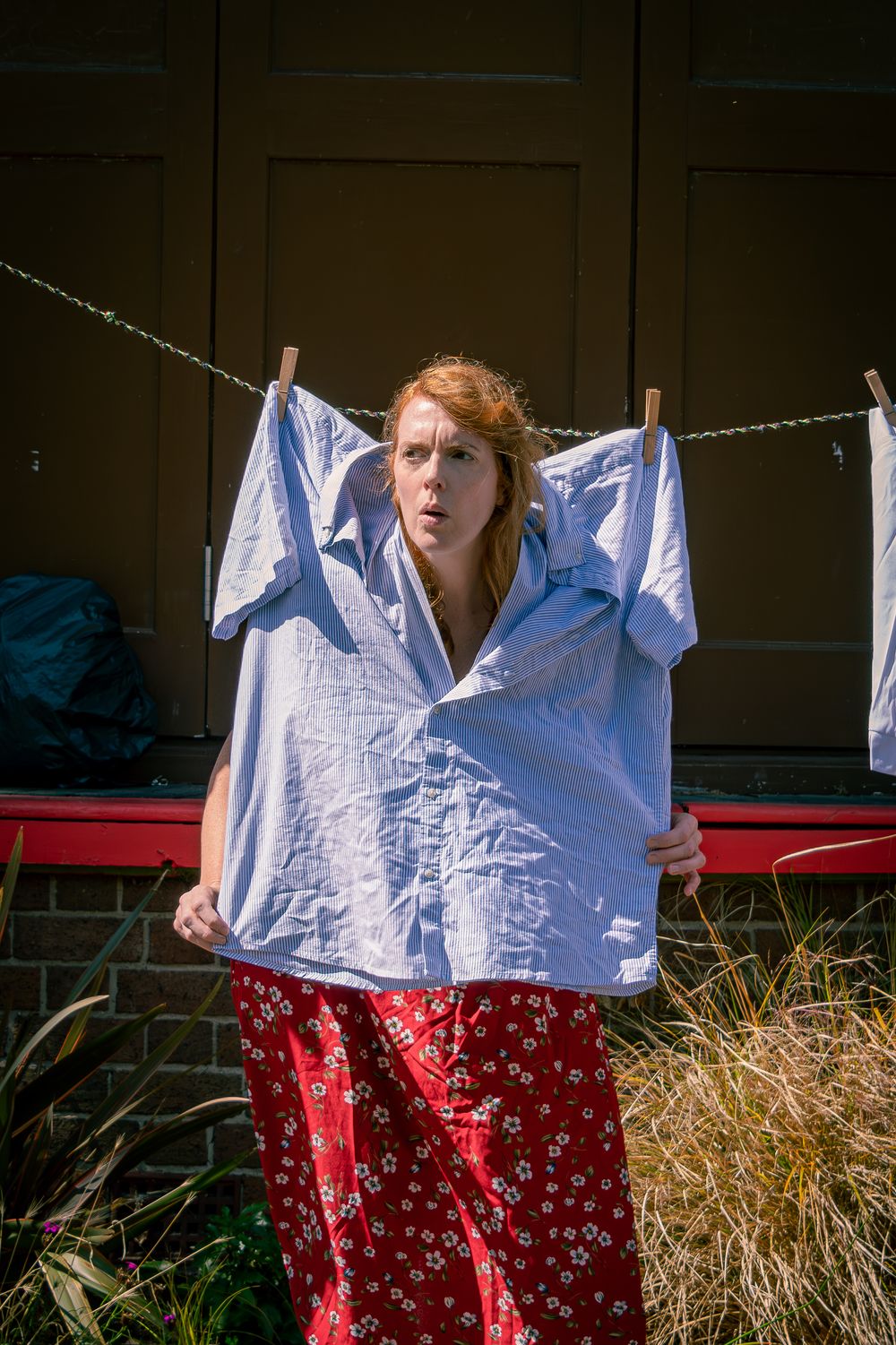 Redhead woman holding large wrinkled shirt near outdoor clothesline with red floral pants.