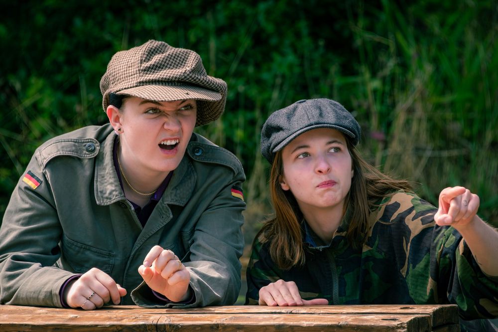 Two young actors in military caps peer over a wooden ledge in an outdoor theatrical scene.