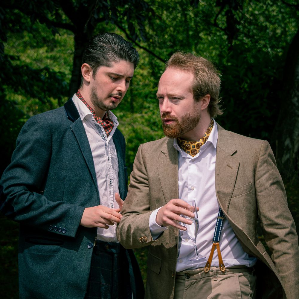 Two men in vintage suits converse outdoors, one wearing a floral cravat, surrounded by lush green trees.