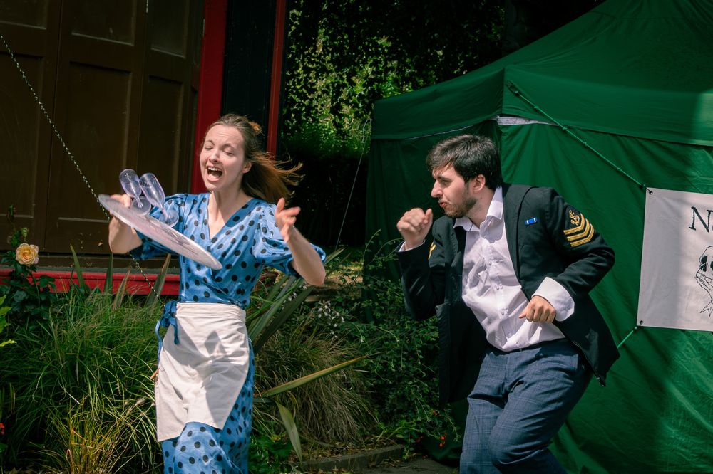 Two actors perform an energetic outdoor theater scene, woman in blue dress and man in uniform reacting dramatically.