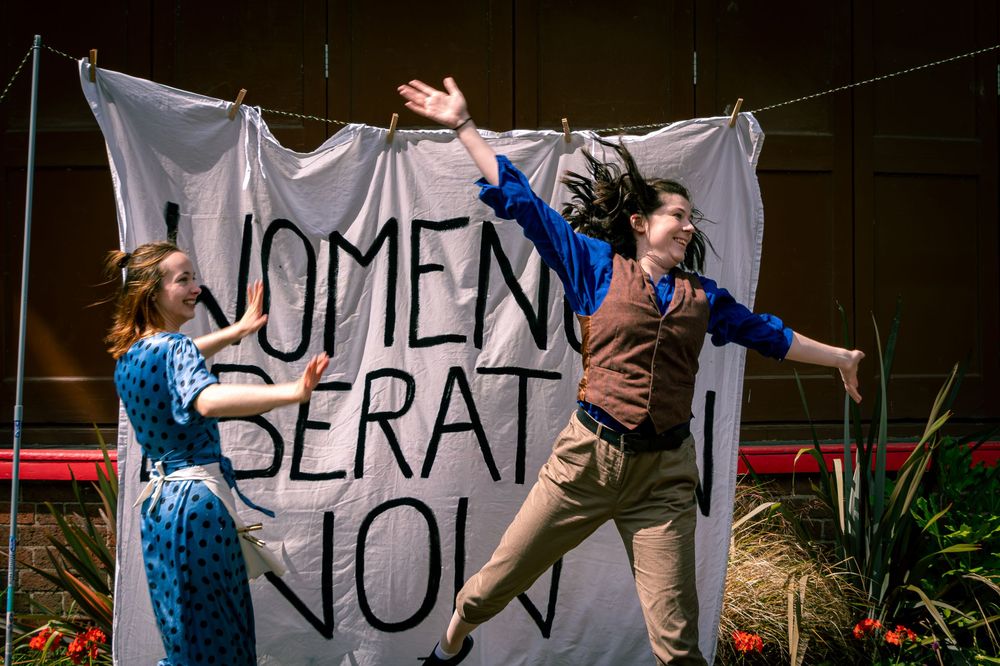 Two performers dance energetically in front of a Women Liberation Now banner on stage.
