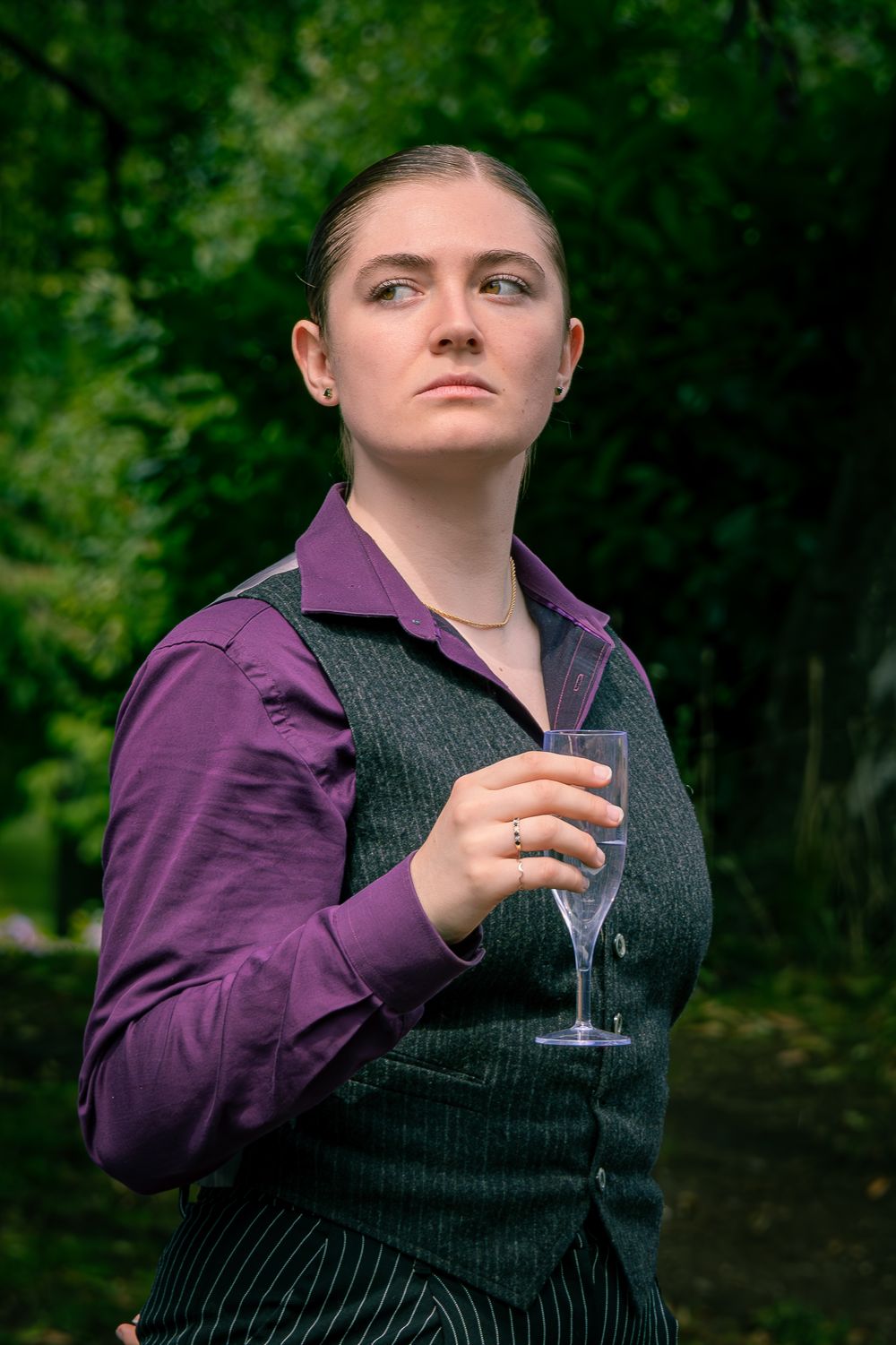 Woman in purple shirt and gray vest holding empty wine glass outdoors with green trees in background.