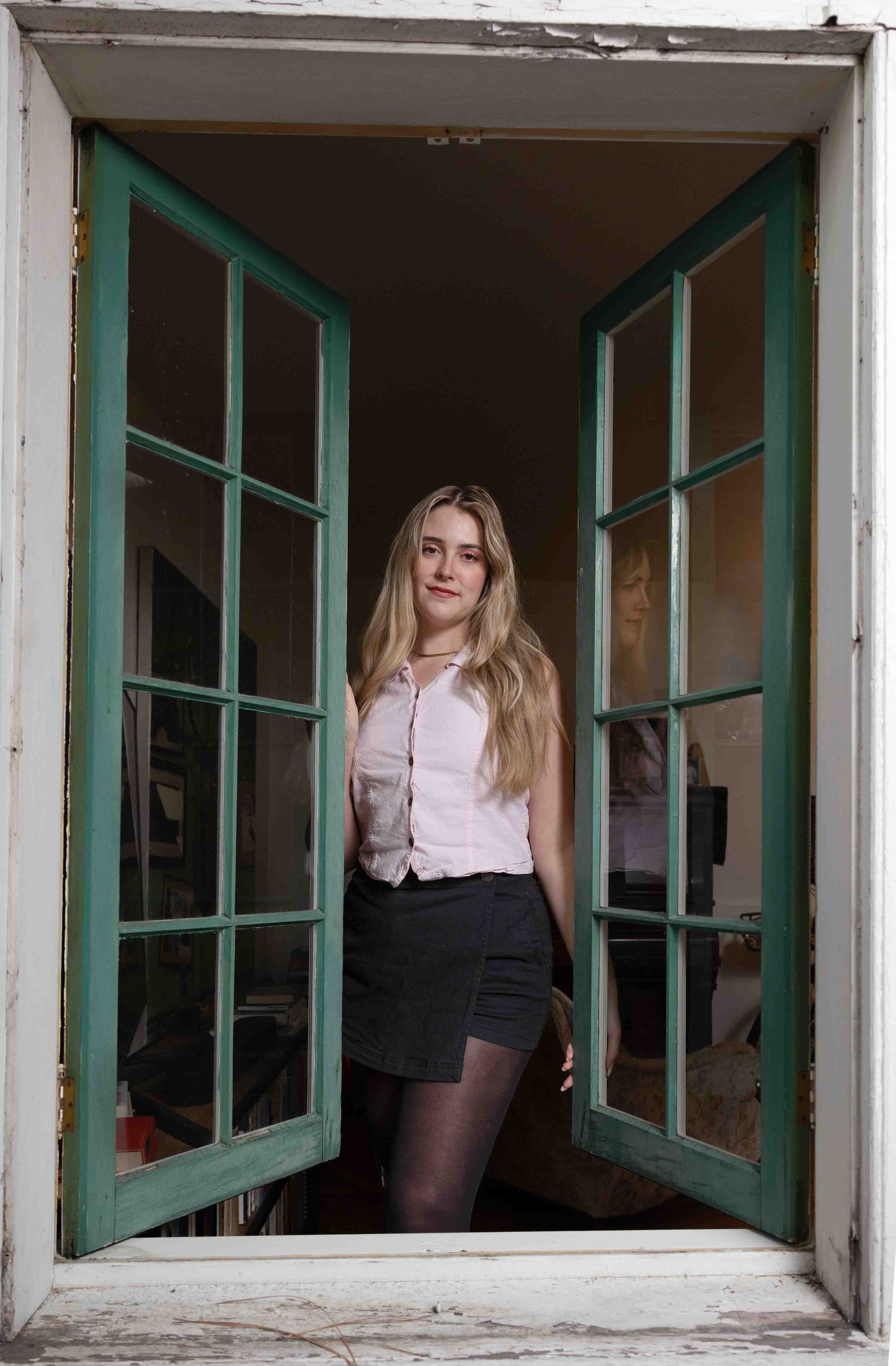 Young blonde woman standing in open green French doors wearing pink blouse and black skirt.