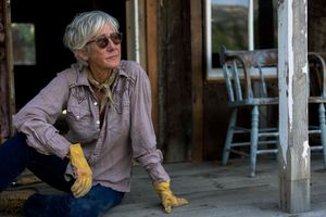 Older woman with gray hair wearing yellow gloves sitting on rustic wooden porch outdoors.