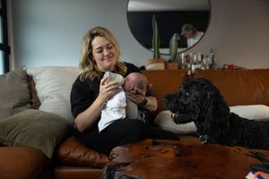 Smiling woman sits on leather sofa bottle-feeding her newborn baby while a black poodle looks on beside her.