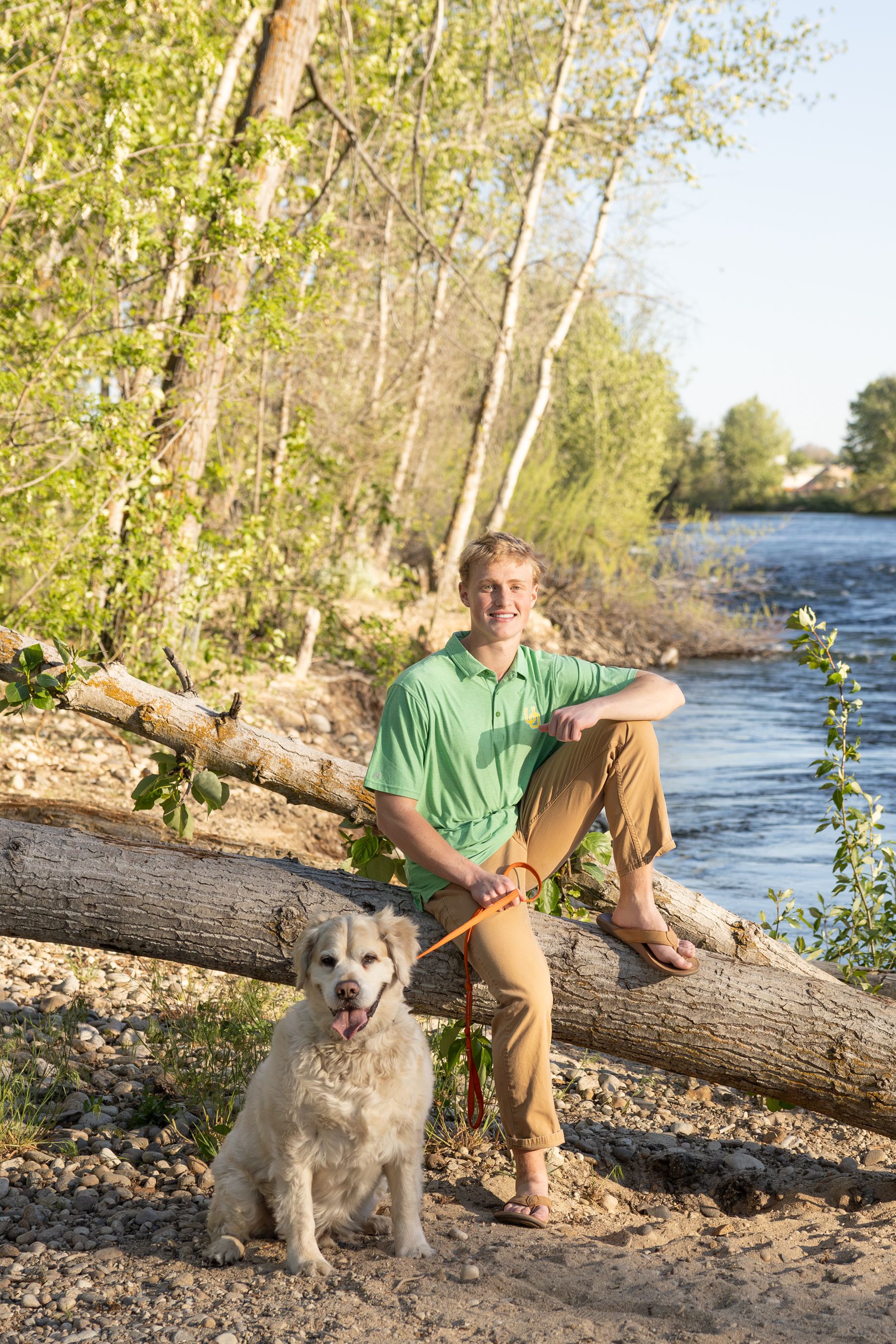 Smiling teenage boy sitting on fallen log with white dog on leash by riverside.