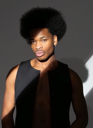Young Black man with natural afro hair wearing a sleeveless black top poses confidently against a gray background.