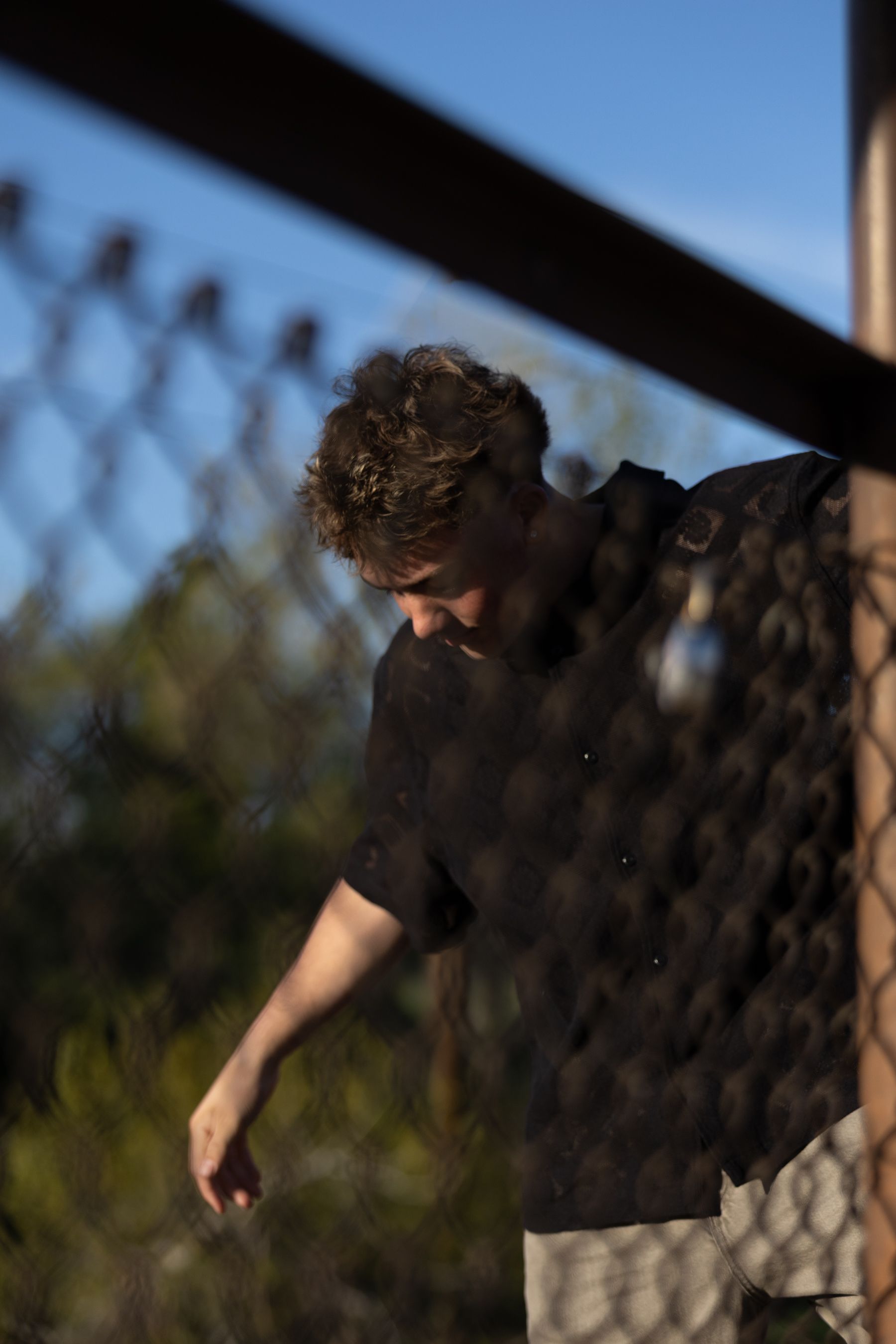 Young man in dark patterned shirt leaning against a chain-link fence in warm golden light.
