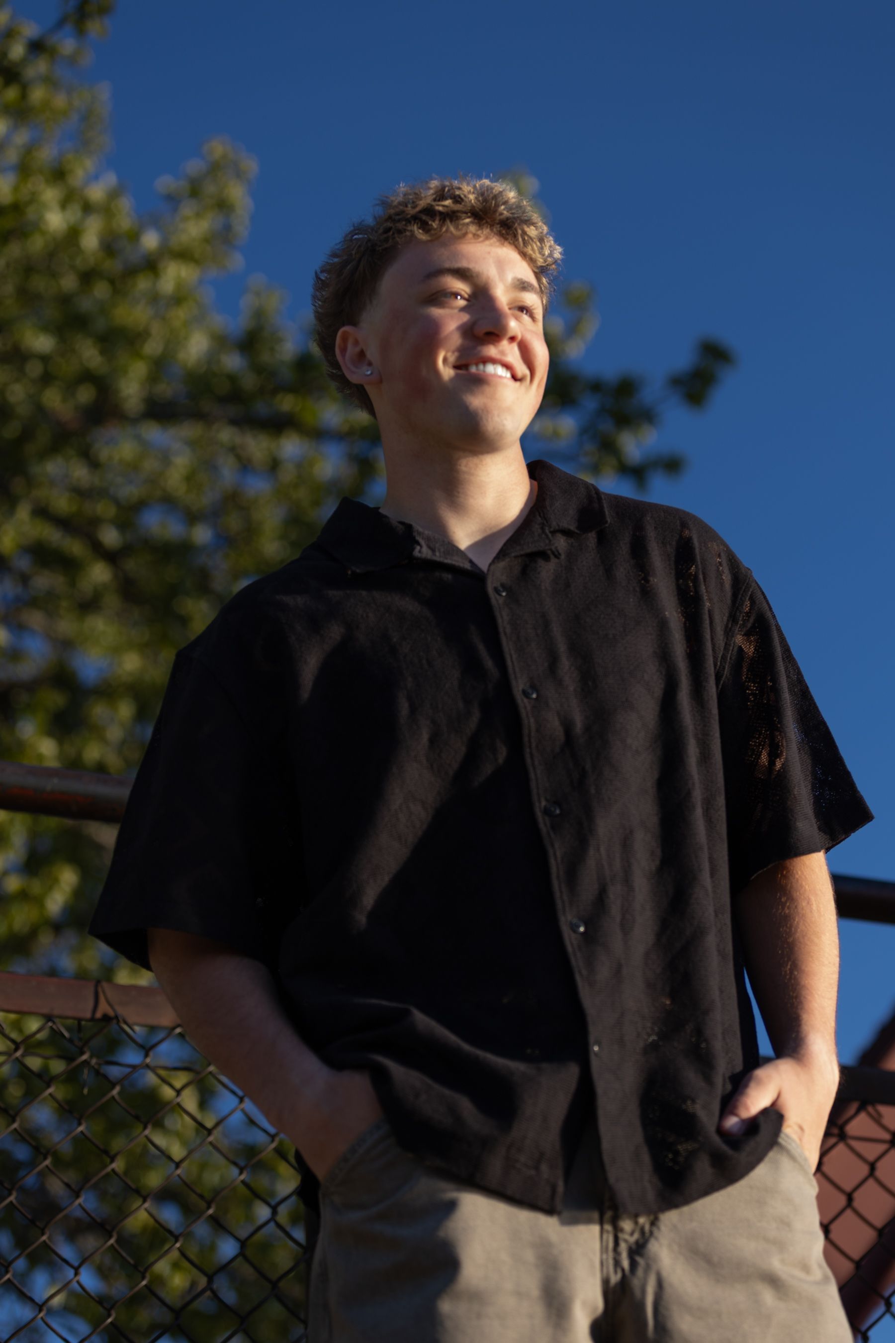 Young man in a black short-sleeve shirt smiling outdoors against a blue sky and chain-link fence.