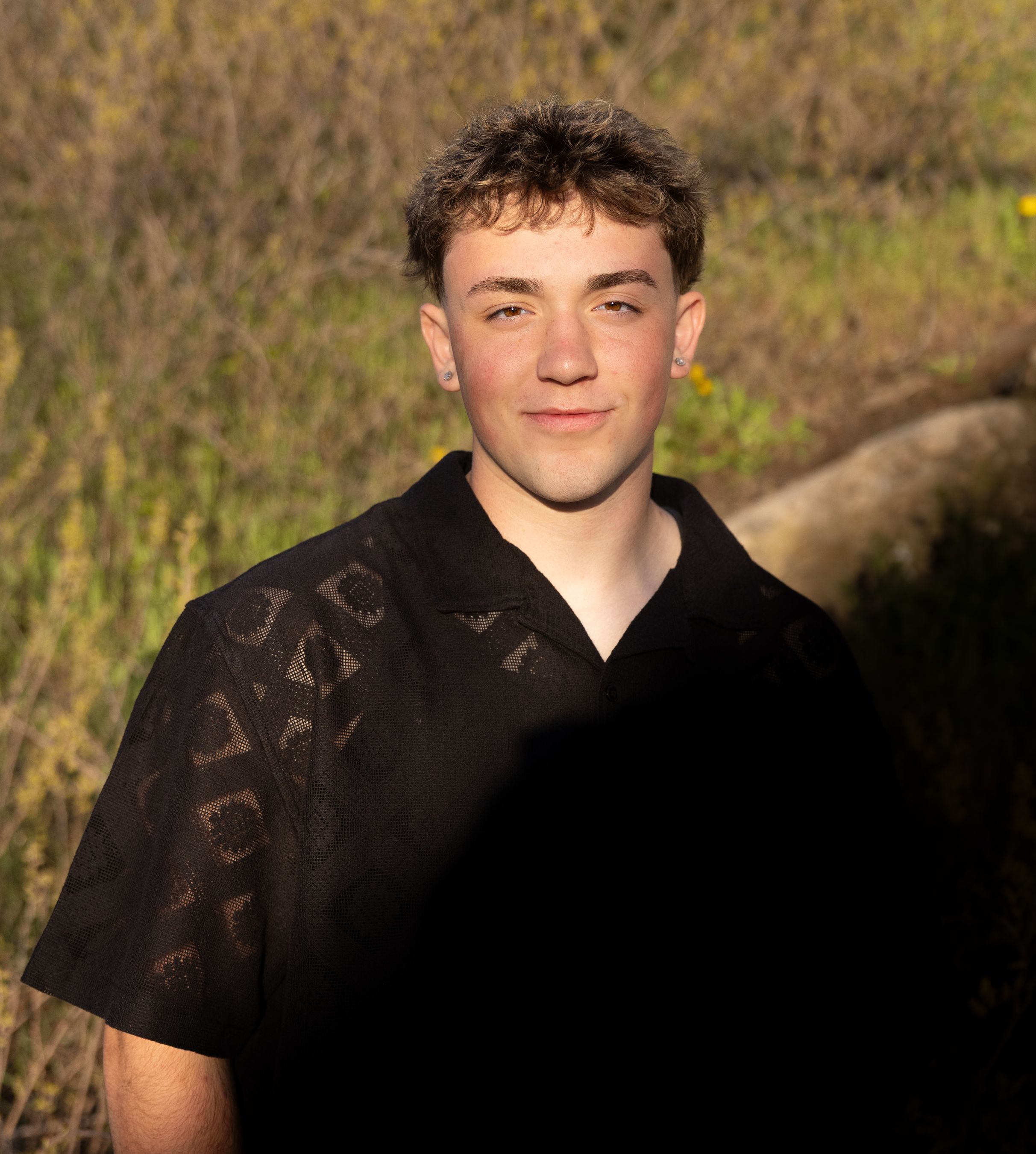 Teen boy in a black patterned shirt posing outdoors in natural sunlight with greenery background.