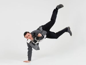 Man in suit balancing on one hand while holding camera in dynamic pose.