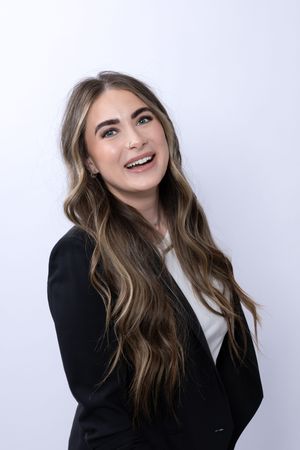Young professional woman with long highlighted hair smiles wearing a black blazer against a white background.