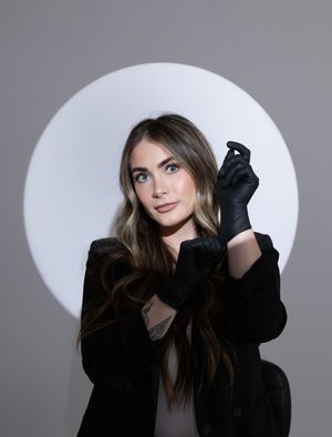 Stylish woman with long highlighted hair wearing black gloves and blazer poses before a circular light backdrop.