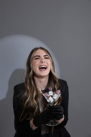 Laughing woman in black blazer and gloves holds a martini glass filled with makeup brushes against gray background.