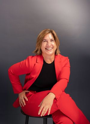 Confident woman in a bold red pantsuit smiles while seated on a stool against a dark gray studio background.
