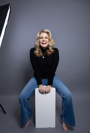 Smiling blonde woman in black turtleneck and flare jeans sitting on white cube in photography studio.