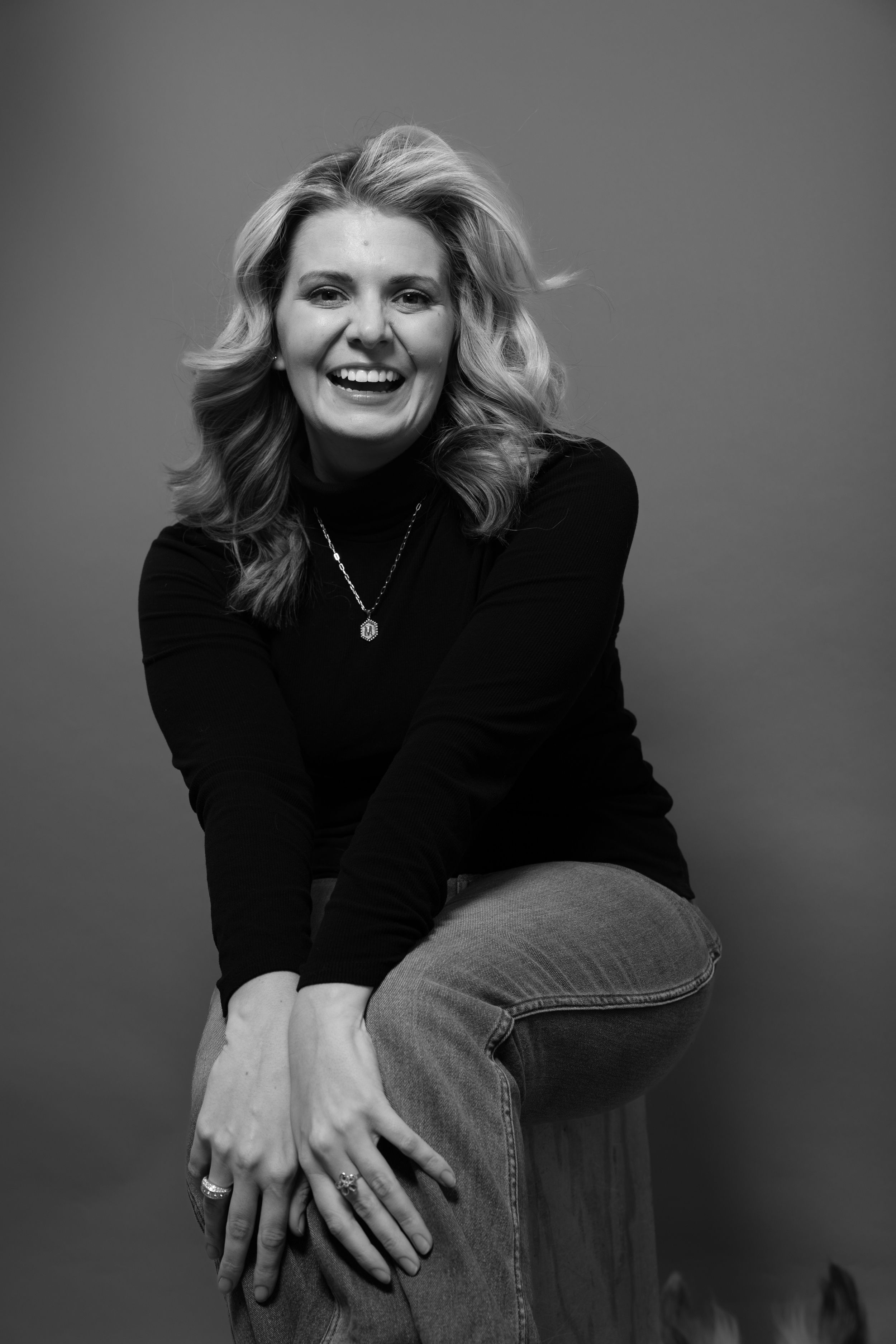 Black and white portrait of laughing blonde woman in black turtleneck and jeans in studio setting.