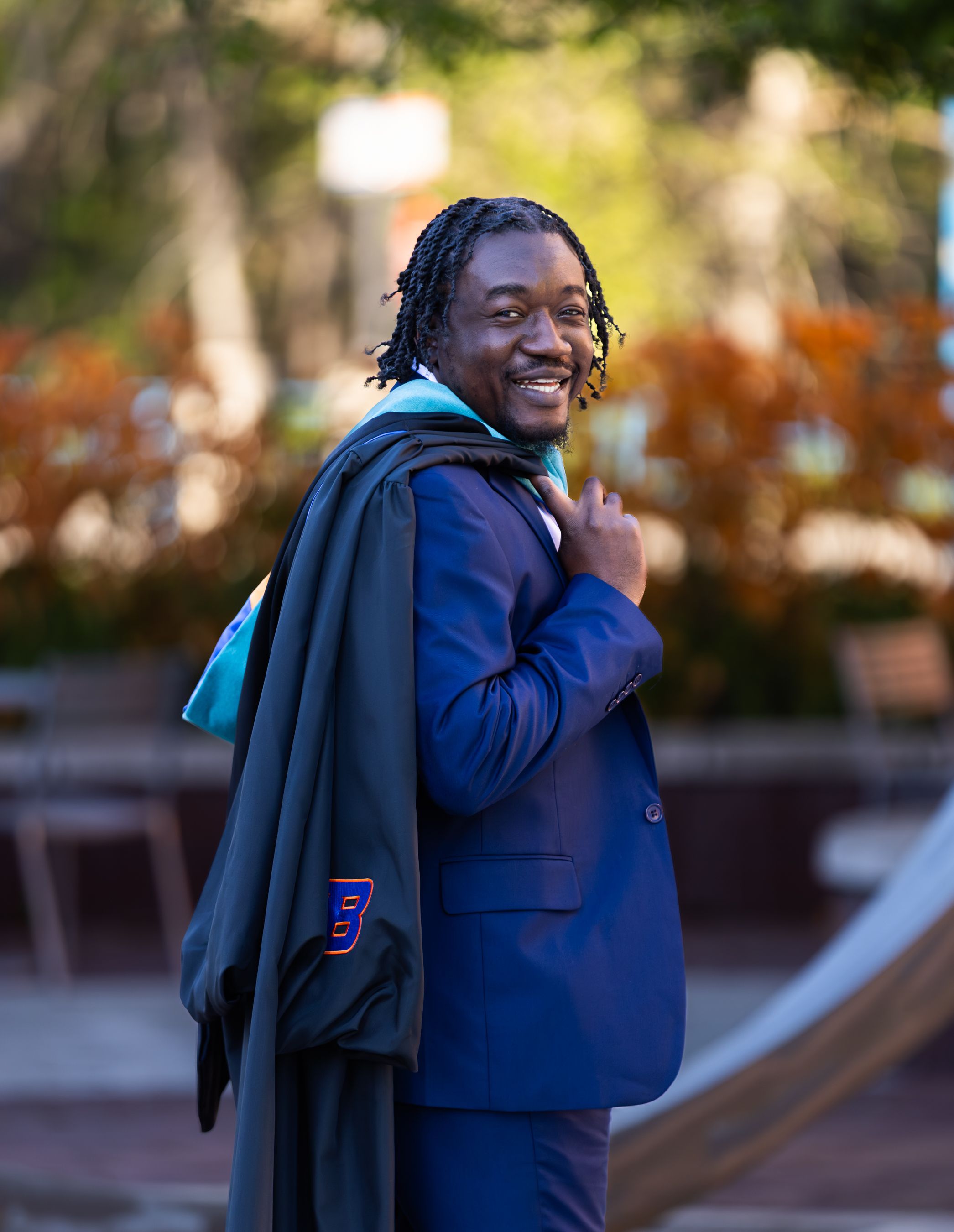 Graduating man in blue suit smiling over shoulder with teal graduation gown on campus.