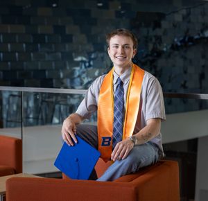 Smiling young man in graduation attire holding blue cap with orange Boise State stole indoors.