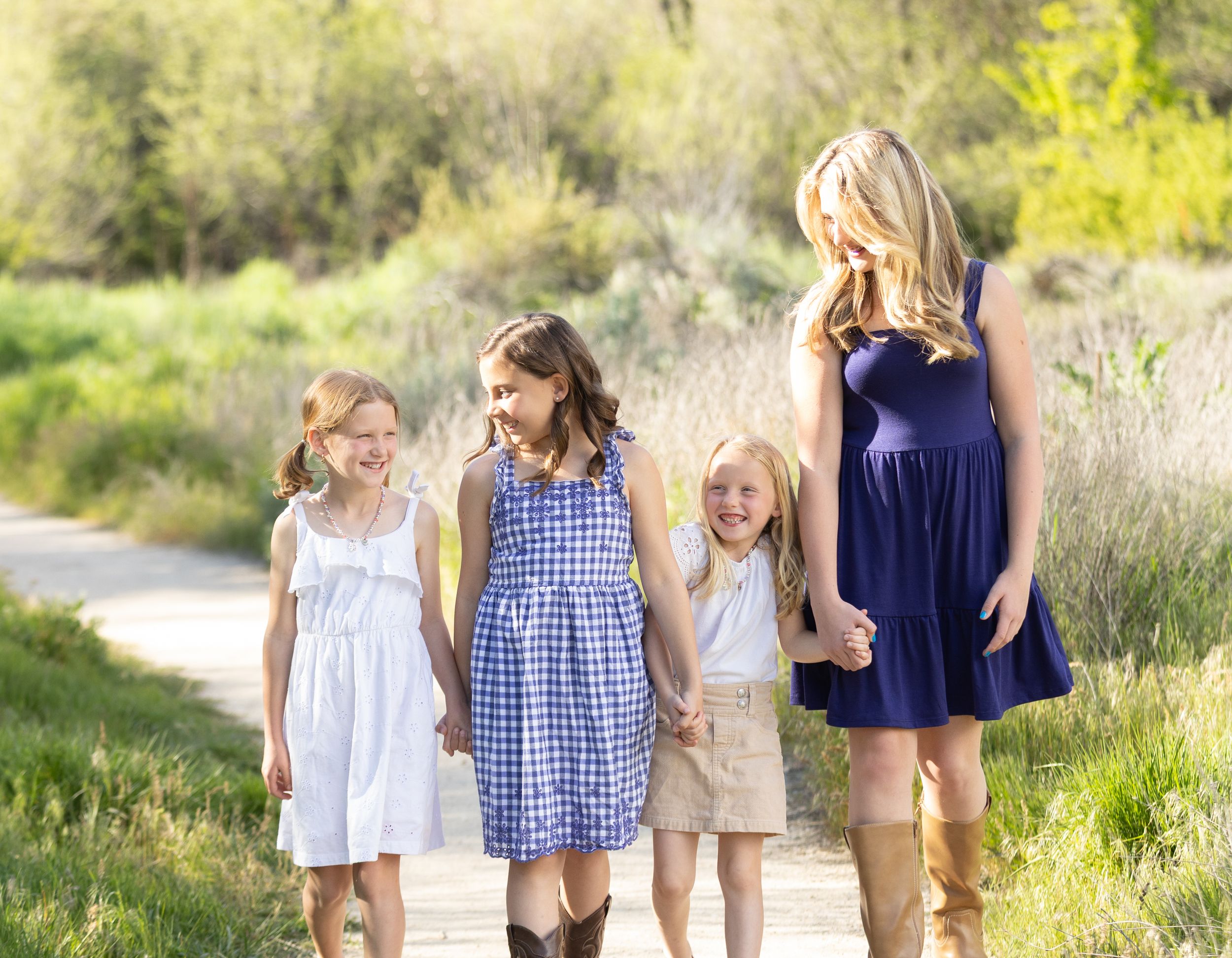 Blonde mother walking with three young daughters on sunny outdoor path surrounded by greenery.