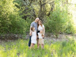 Happy family of four poses together in a lush green meadow surrounded by trees during a outdoor photo session.