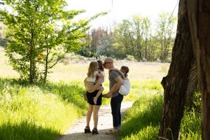 Family of four sharing kisses and hugs on a sunny nature path surrounded by vibrant green grass and trees.