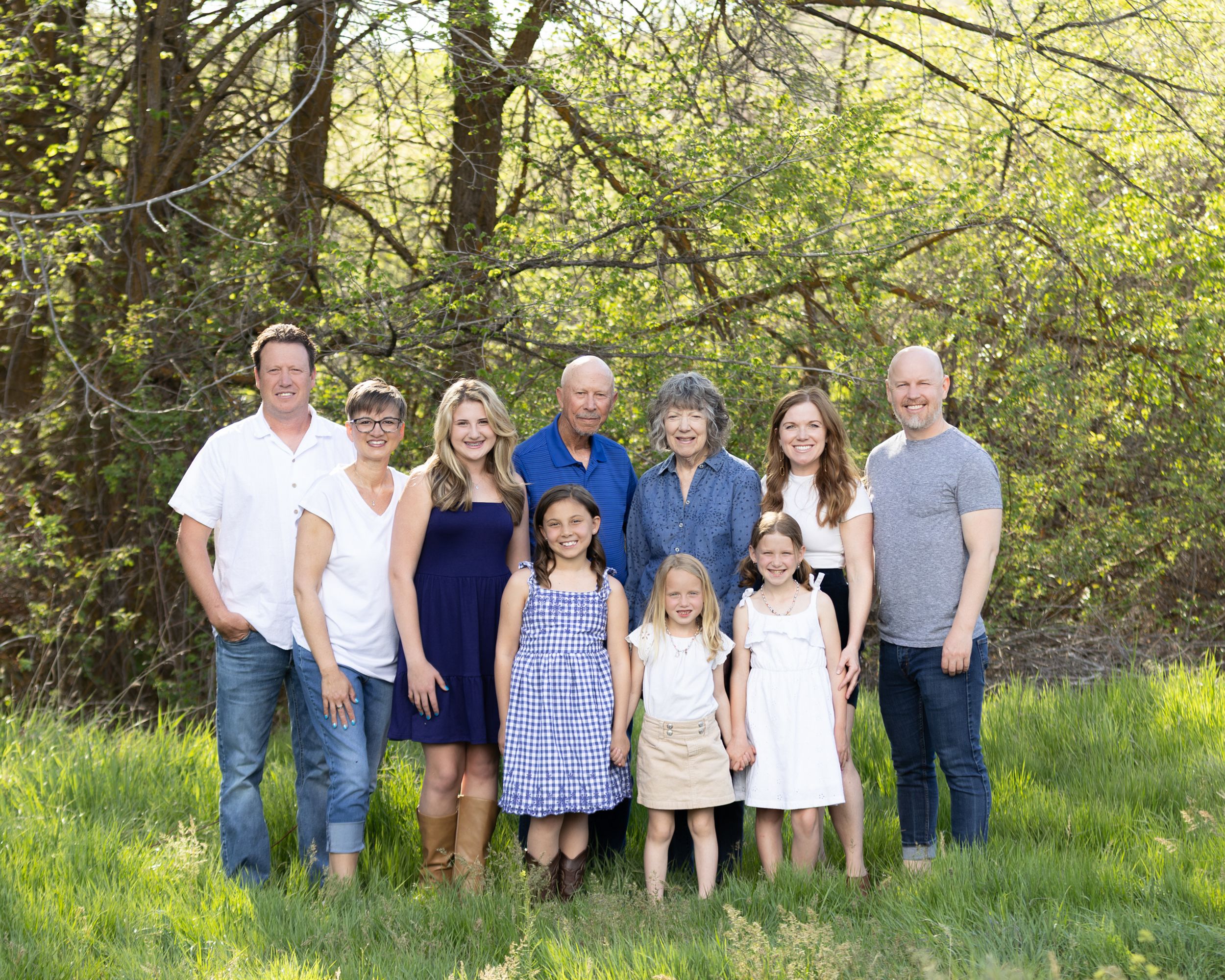 Multi-generational family portrait outdoors in a lush green spring setting with trees in background.