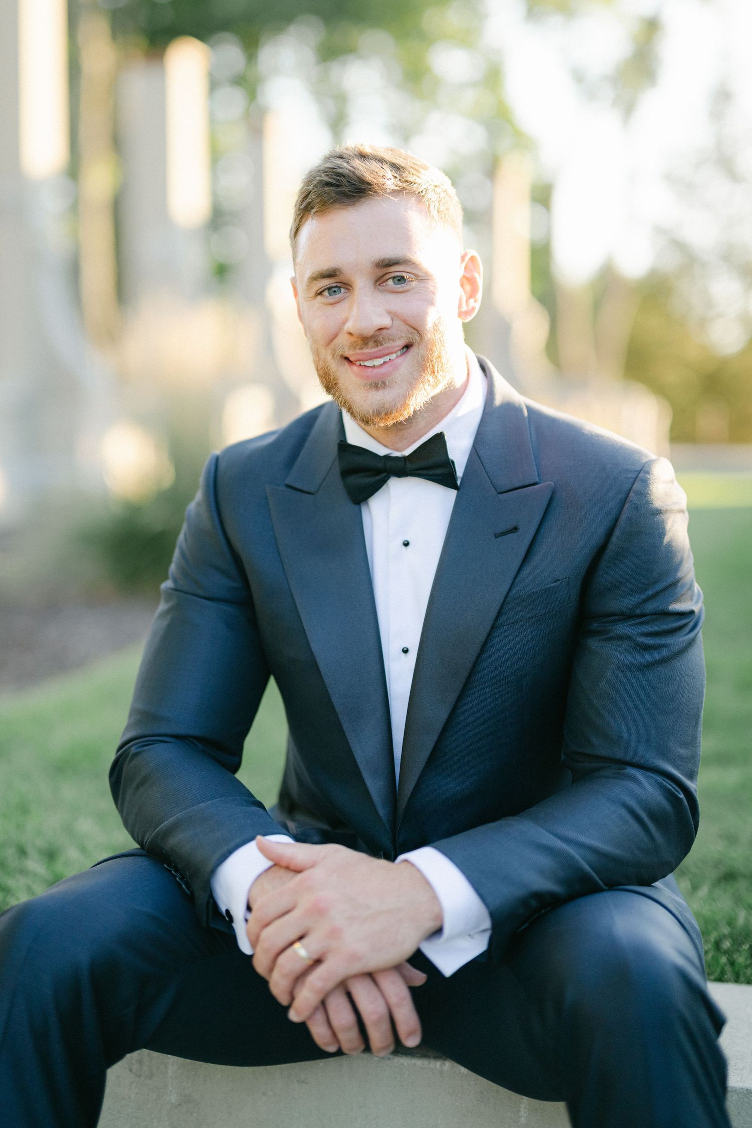 Armour House Wedding - Smiling groom in black tuxedo and bow tie sits outdoors on stone ledge with hands clasped.
