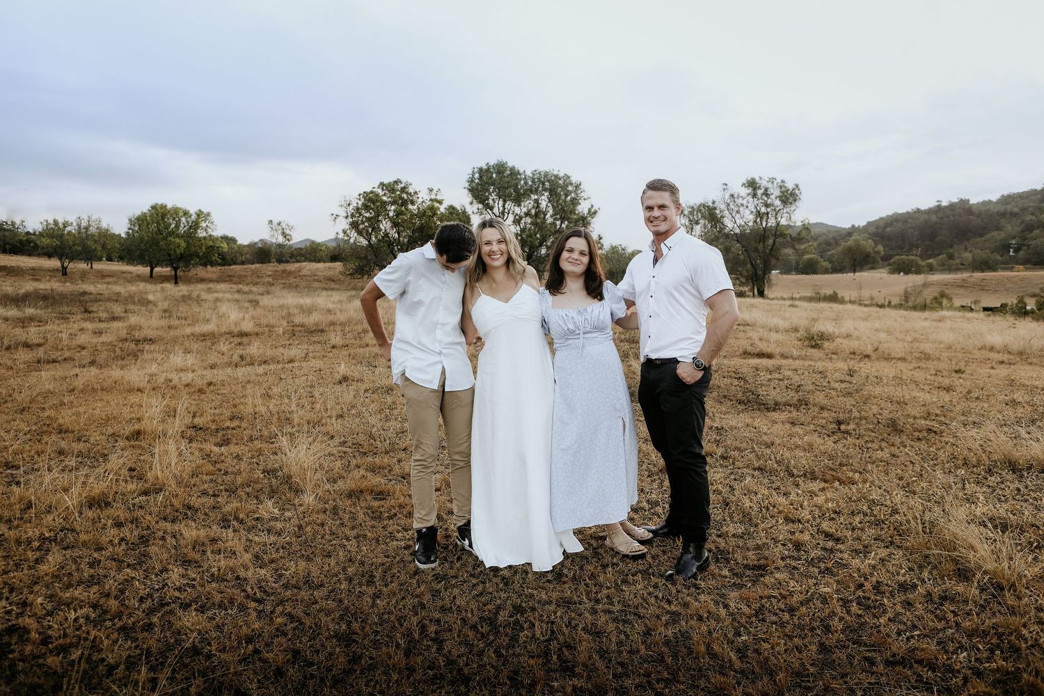 Four people in white outfits posing together in a golden field with trees and overcast sky in the background.