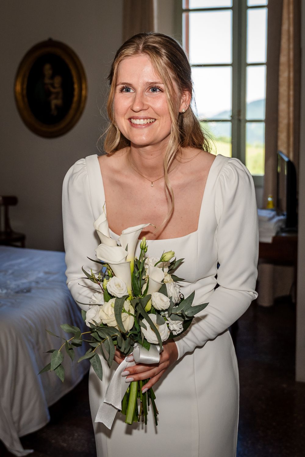 Smiling bride in white long-sleeve gown holding elegant bouquet of white roses and greenery indoors.