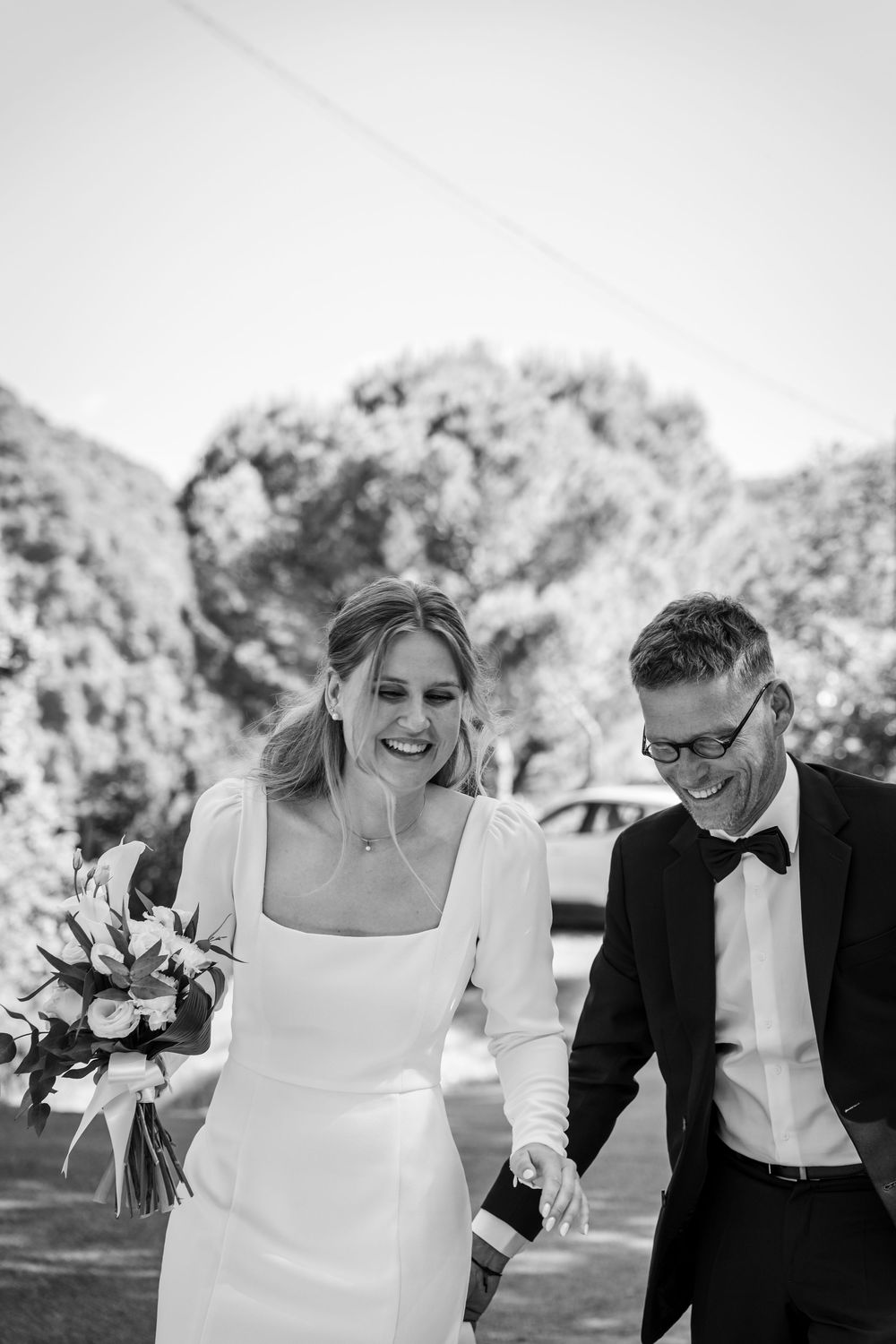Black and white photo of a laughing bride holding bouquet walking with groom in bow tie on wedding day.