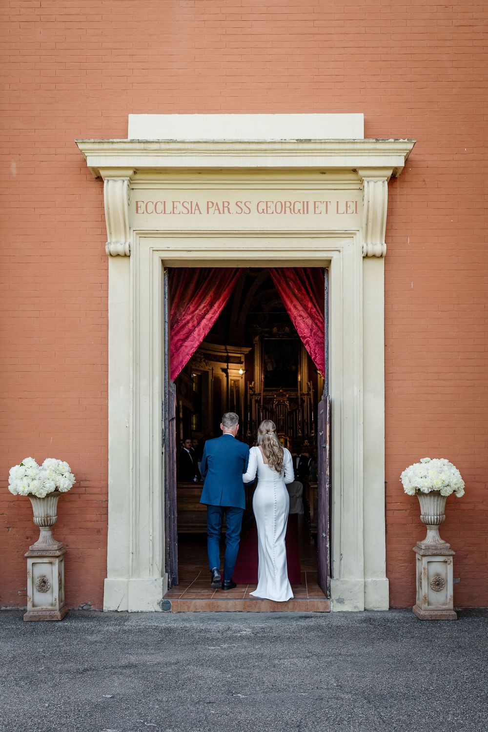 Bride and groom entering an Italian church through ornate doors flanked by white floral arrangements.