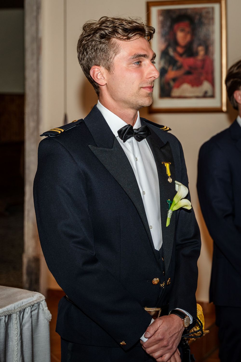 Young man in military dress uniform with bow tie and white flower boutonniere standing at formal event.