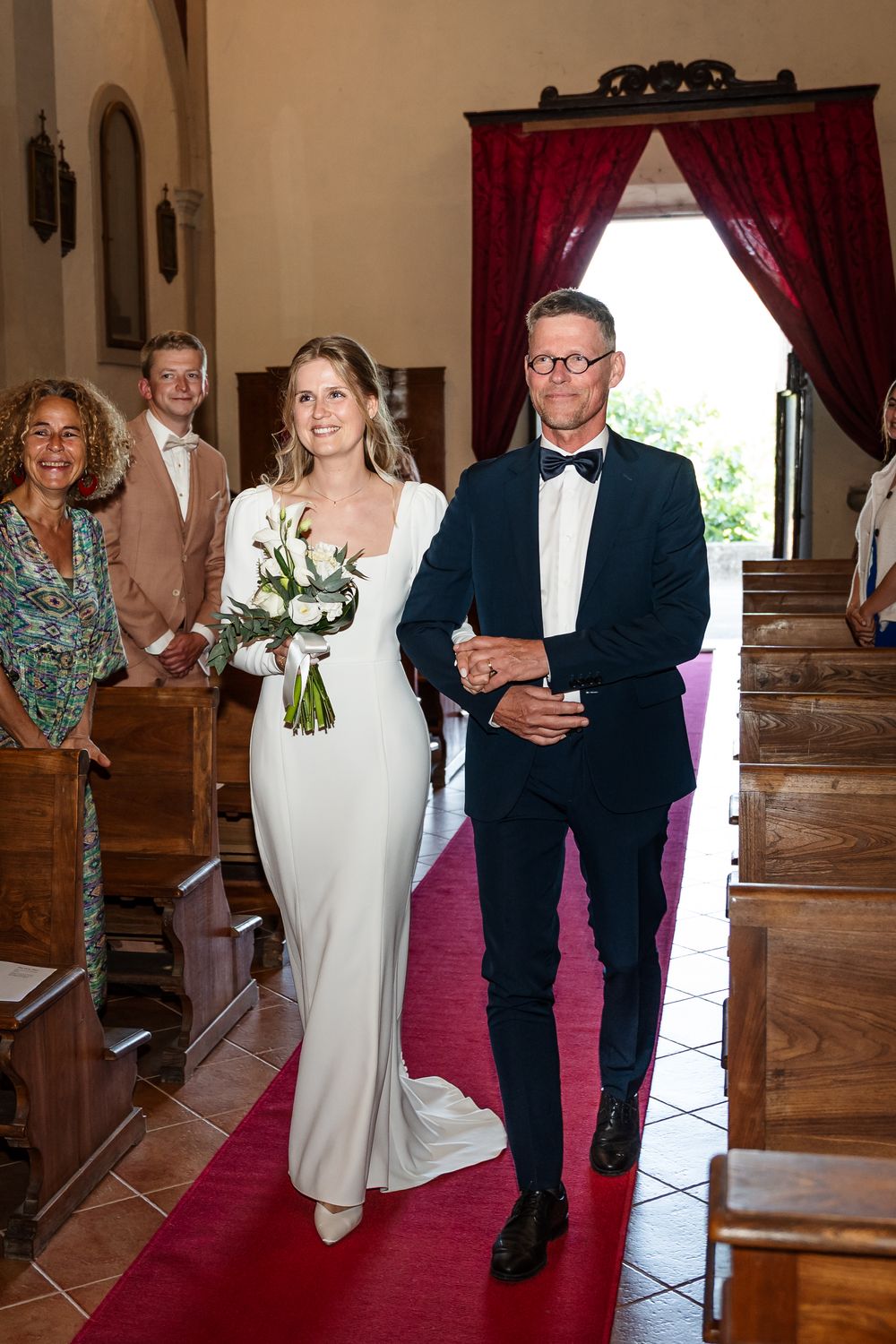 Bride in white gown walks down red carpet aisle with escort in navy tuxedo during church wedding ceremony.
