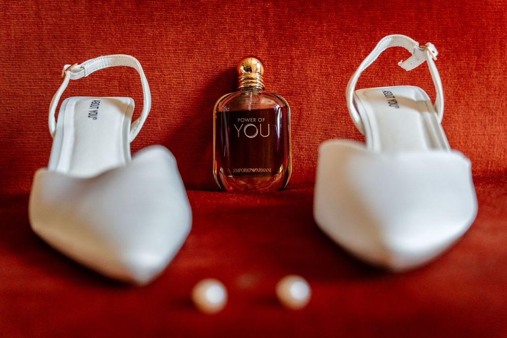 White bridal heels and pearl earrings flanking an Emporio Armani YOU perfume bottle on red velvet.