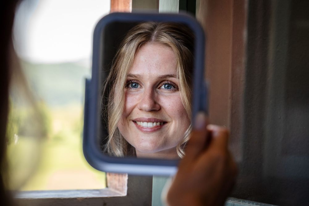Smiling blonde bride with blue eyes reflected in a small handheld mirror on wedding day.