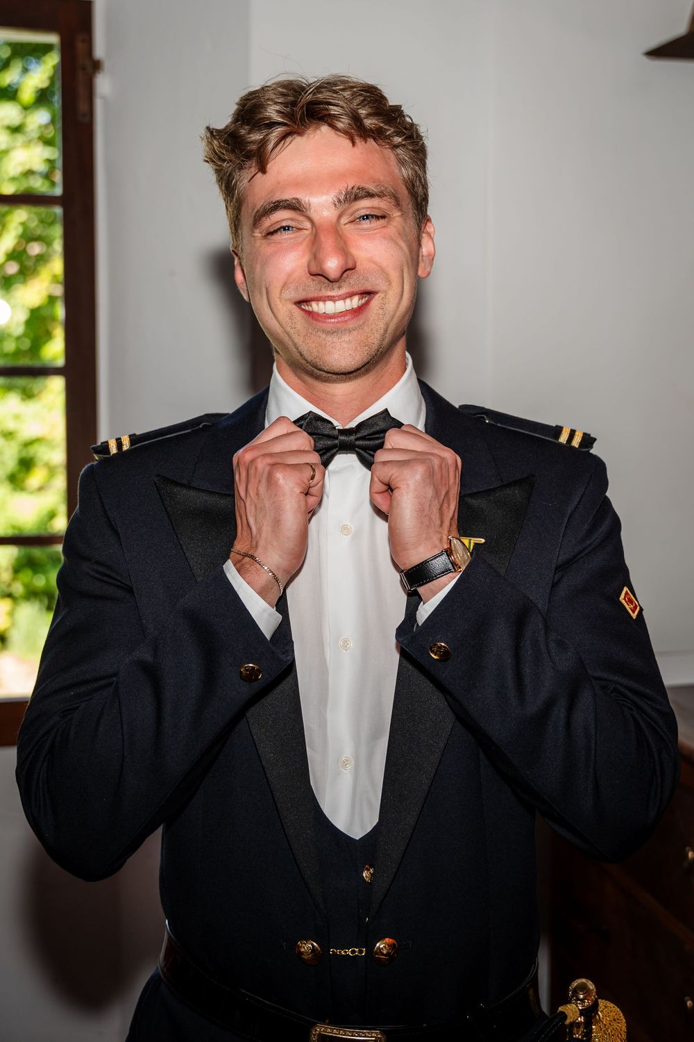 Smiling man in military dress uniform adjusting black bow tie, wearing gold-buttoned jacket with epaulettes.