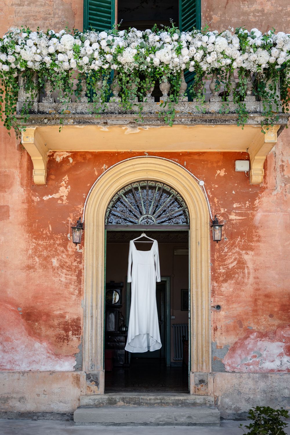 White wedding dress hanging in an arched doorway of a rustic Italian villa with floral balcony above.