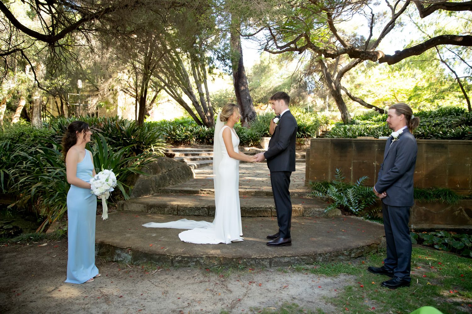 Wedding couple kissing surrounded by smiling family members in elegant attire outdoors.