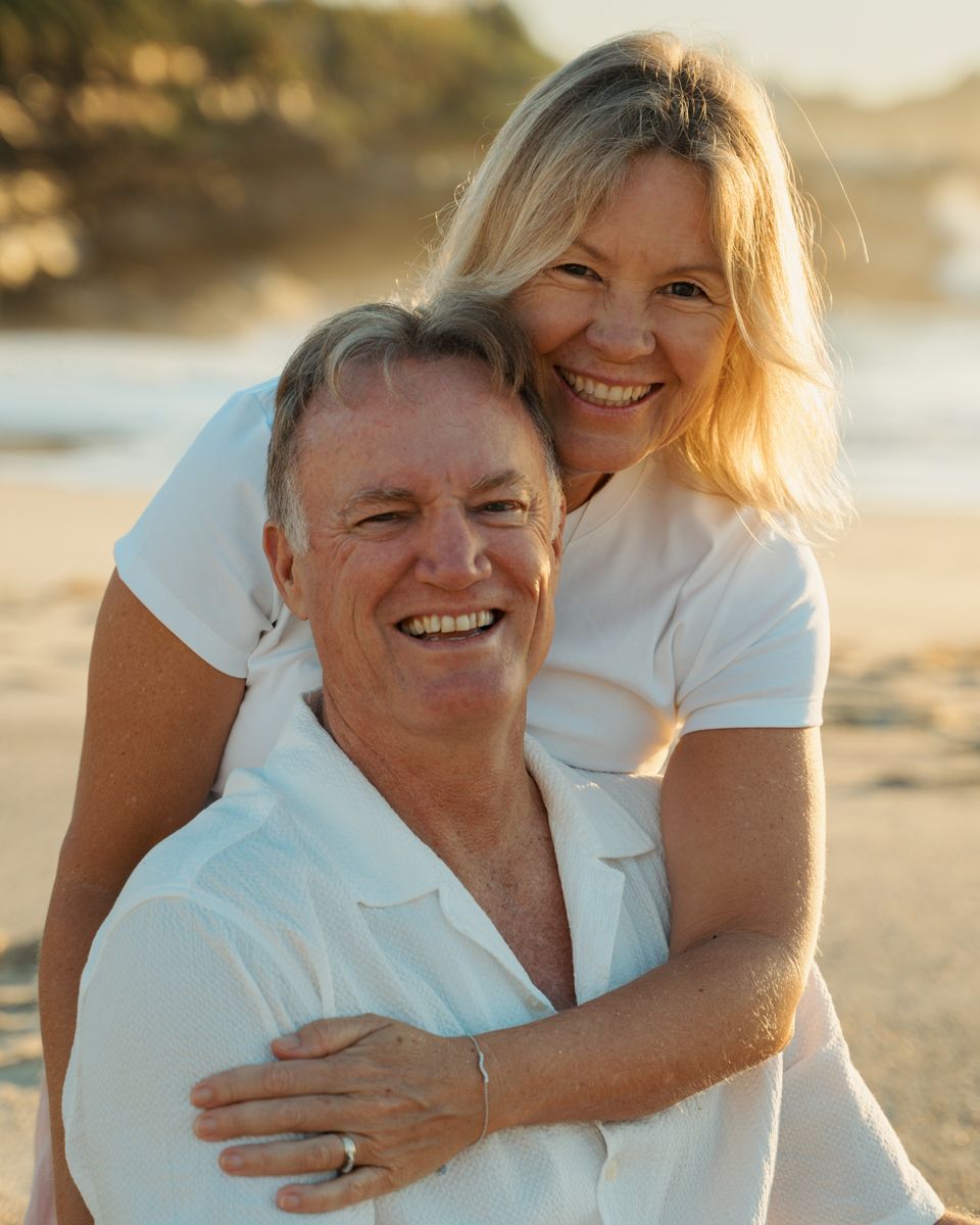 Happy couple on a sunny beach, both wearing white, smiling warmly with golden light and ocean in the background Bronte Phototoshoot