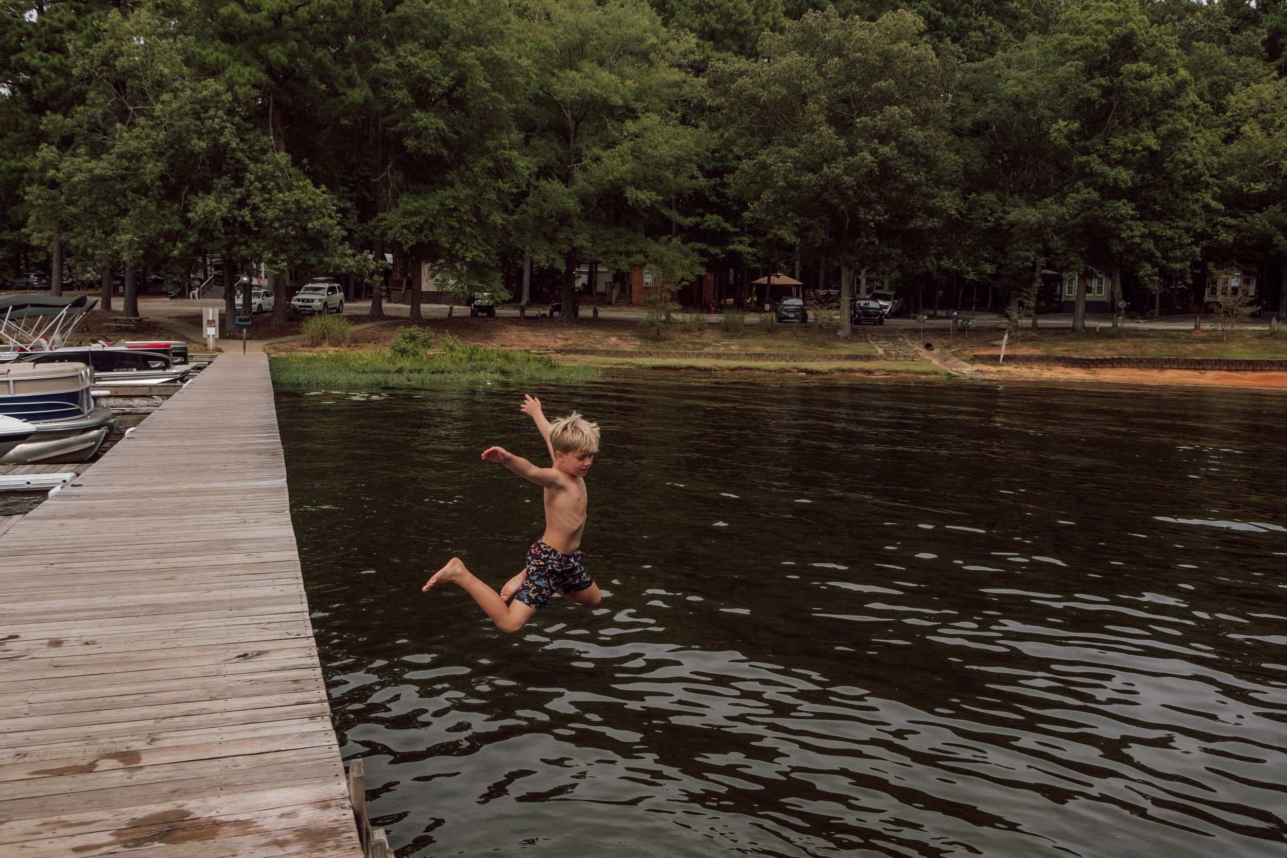 A child leaps off a wooden dock into a calm lake surrounded by lush green trees on a summer day hampton roads documentary family photography