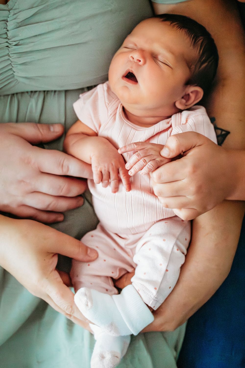 Newborn baby in pink onesie yawning while being held by parents hands.