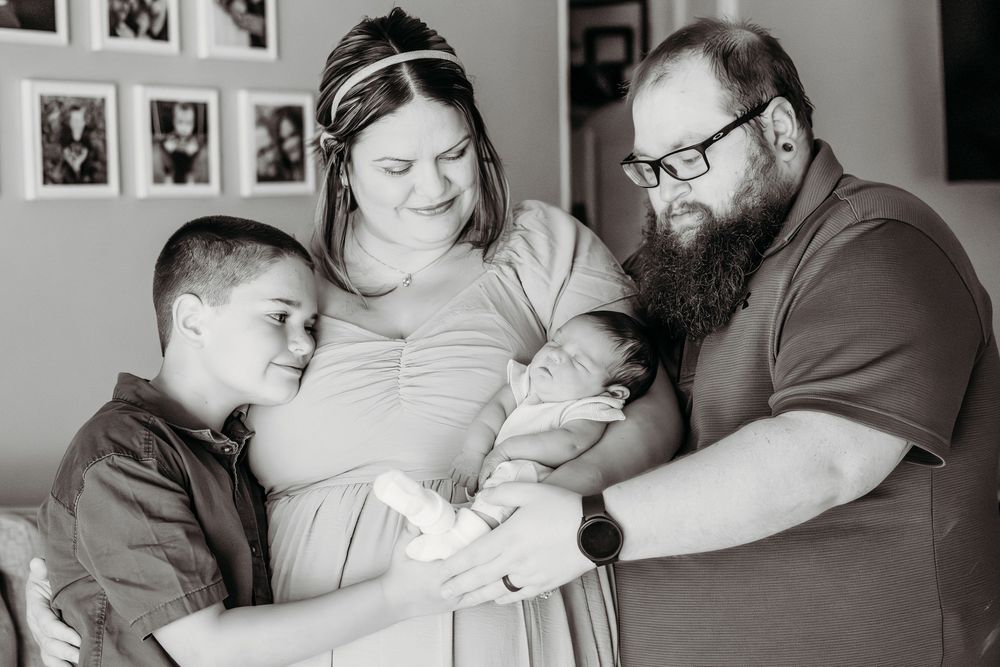Black and white family photo of parents with newborn baby and young son in a cozy home setting.