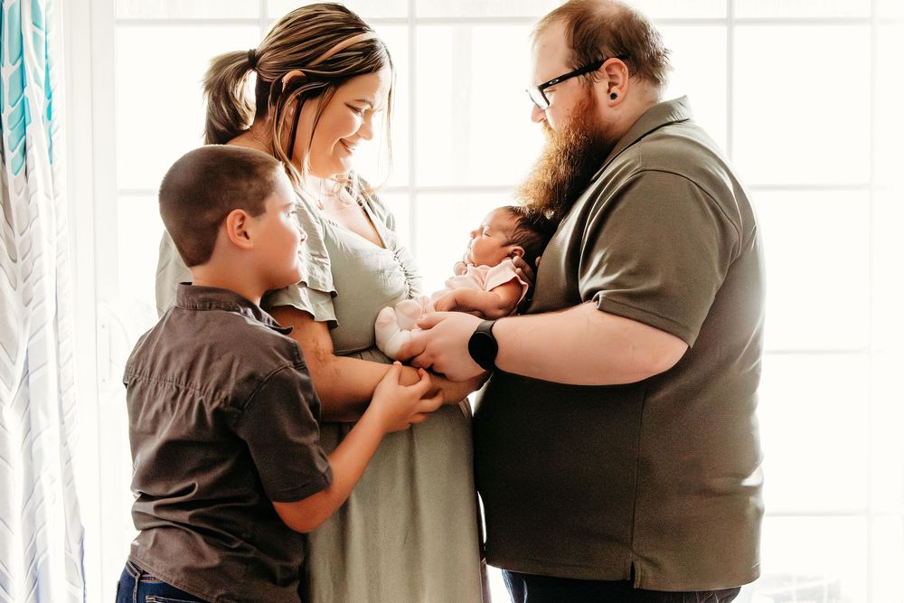 Family of four admiring their newborn baby together near a bright window in a warm, intimate moment.