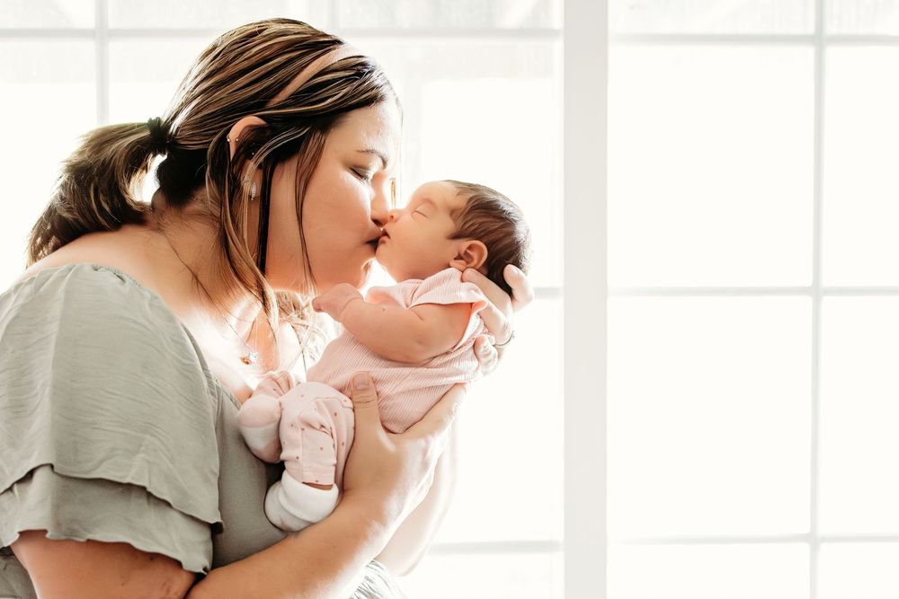 A mother gently kissing her newborn baby in a softly lit, tender portrait.
