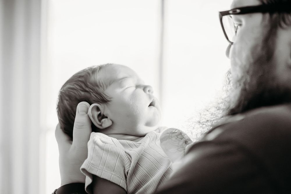 A bearded father tenderly cradles his sleeping newborn baby in a black and white portrait photo.