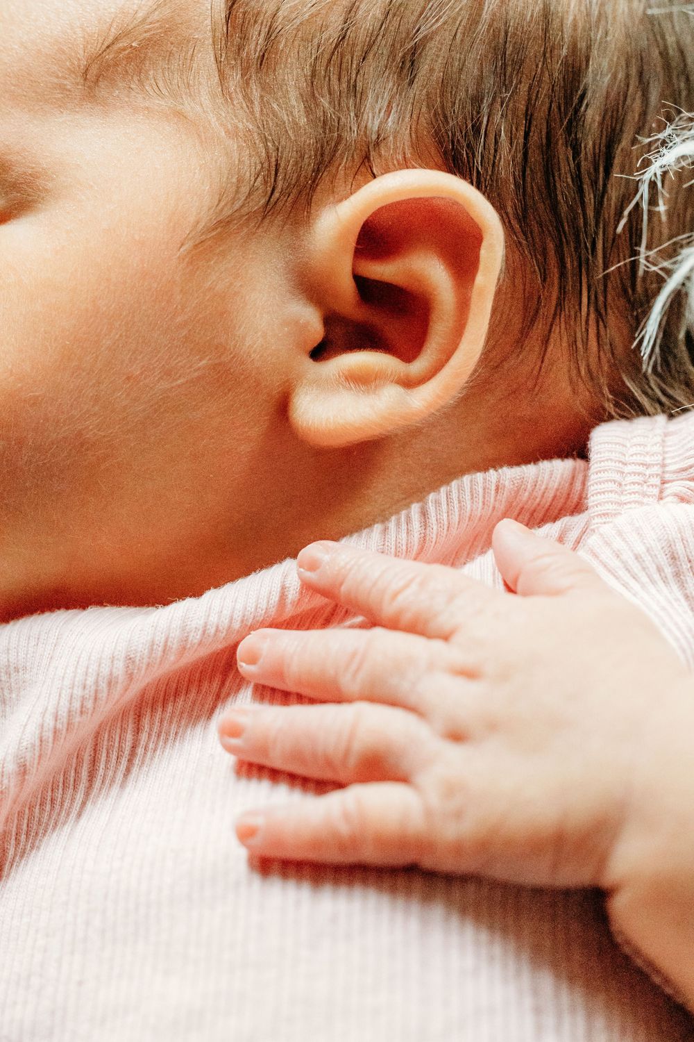 Close-up of a sleeping newborn baby's ear and tiny hand resting on a pink ribbed onesie.