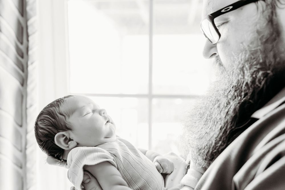 Black and white photo of a bearded man tenderly holding a sleeping newborn baby near a bright window.