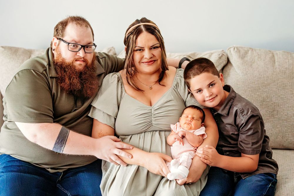 Happy family of four posing on a couch, parents holding a newborn baby with an older child smiling beside them.